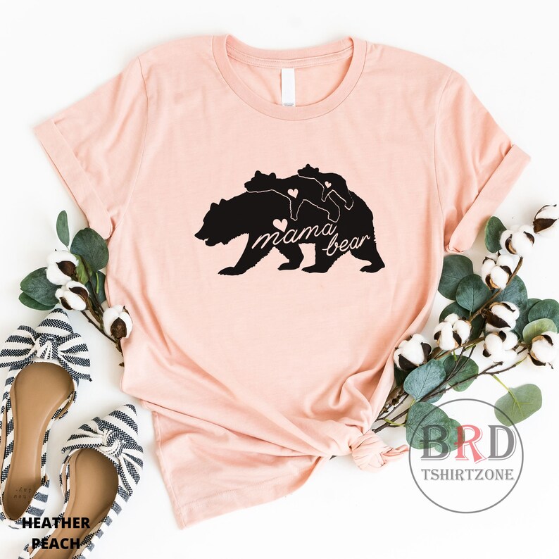 May include: Heather peach-colored t-shirt featuring a black graphic of a mama bear with two cubs and the words "mama bear". The design includes heart accents. The shirt is styled with flats and greenery.