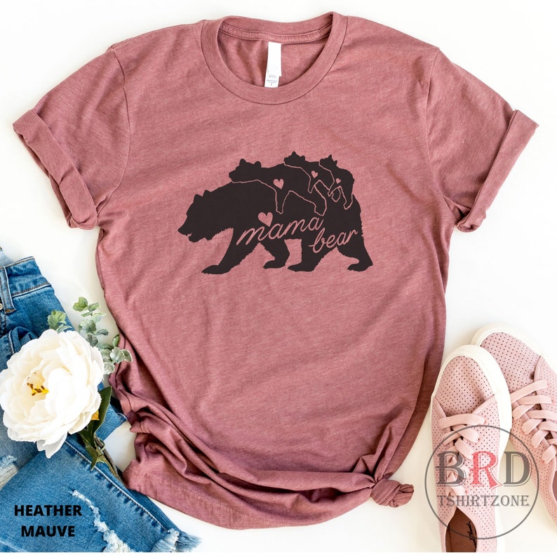 May include: Heather mauve t-shirt featuring a black graphic of a mama bear with three cubs and the text "mama bear". The shirt is folded, with a pair of pink shoes and denim jeans visible in the background. The shirt is made of a soft material.