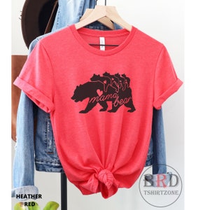 May include: Heather red t-shirt with a black graphic of a bear and cubs, with the words "mama bear" in a script font. The shirt is displayed on a hanger, with a denim jacket and hat in the background. The shirt is knotted at the bottom.