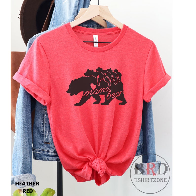 May include: Heather red t-shirt with a black graphic of a bear and cubs, with the words "mama bear" in a script font. The shirt is displayed on a hanger, with a denim jacket and hat in the background. The shirt is knotted at the bottom.