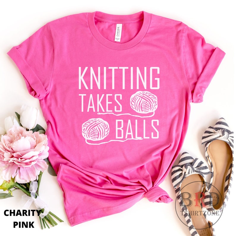 May include: Pink t-shirt with the text "Knitting Takes Balls" in white lettering. Two white yarn balls are depicted on the shirt.
