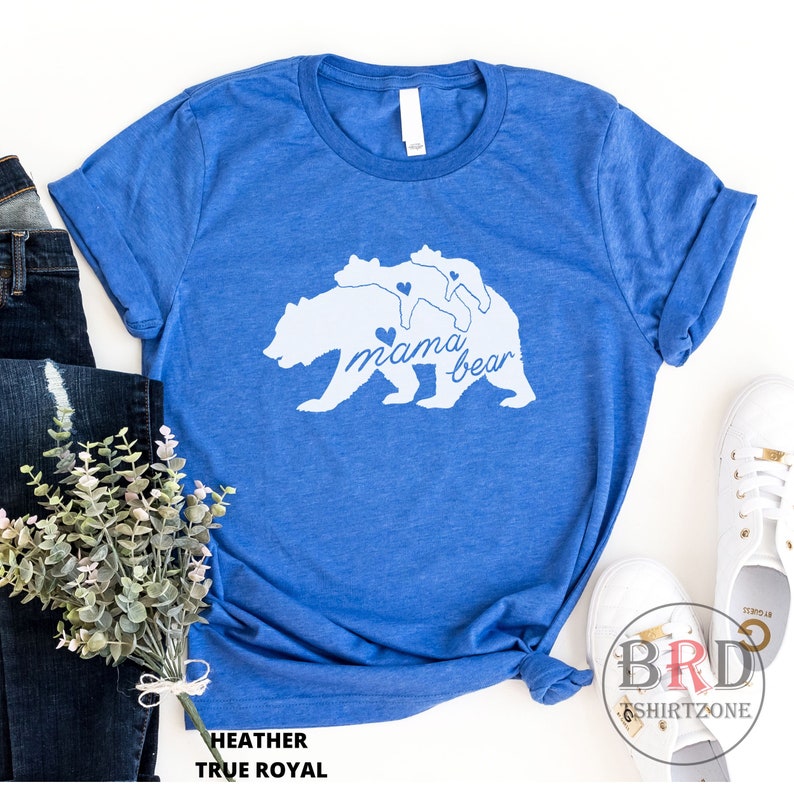 May include: A blue heather t-shirt with a white graphic of a mama bear and two cubs, with the words "mama bear" in cursive. The shirt is styled with rolled-up sleeves and a knot at the bottom. The text "HEATHER TRUE ROYAL" is at the bottom.