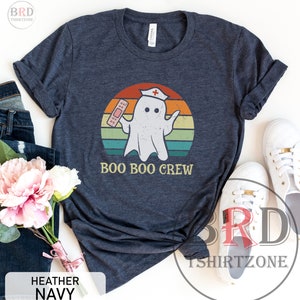 May include: Heather navy t-shirt with a distressed graphic of a ghost wearing a nurse's cap and holding a bandage. The text "BOO BOO CREW" is below the ghost, set against a retro sunset design.
