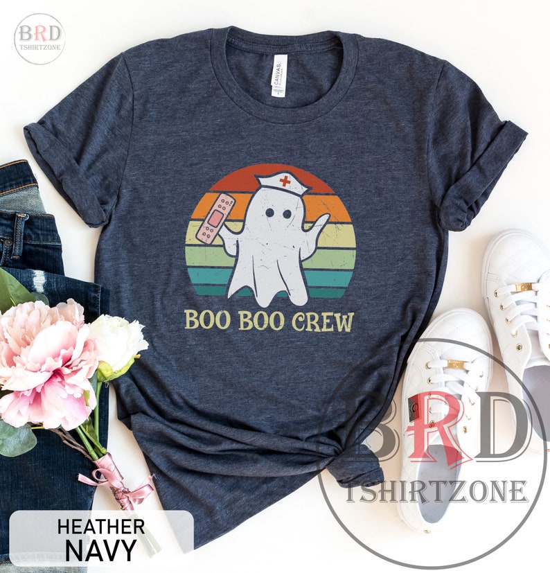 May include: Heather navy t-shirt with a distressed graphic of a ghost wearing a nurse's cap and holding a bandage. The text "BOO BOO CREW" is below the ghost, set against a retro sunset design.