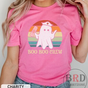 May include: A pink t-shirt with the text "BOO BOO CREW" and a cartoon ghost wearing a nurse's hat and holding a bandage. The ghost is set against a retro sunset design with orange, yellow, green, and blue stripes.
