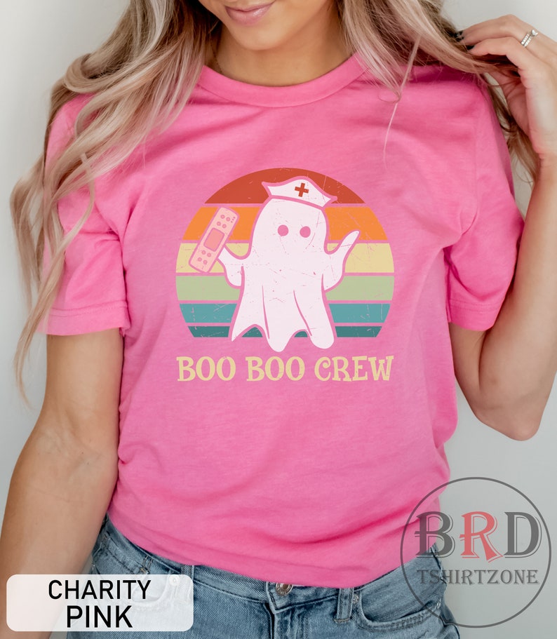 May include: A pink t-shirt with the text "BOO BOO CREW" and a cartoon ghost wearing a nurse's hat and holding a bandage. The ghost is set against a retro sunset design with orange, yellow, green, and blue stripes.