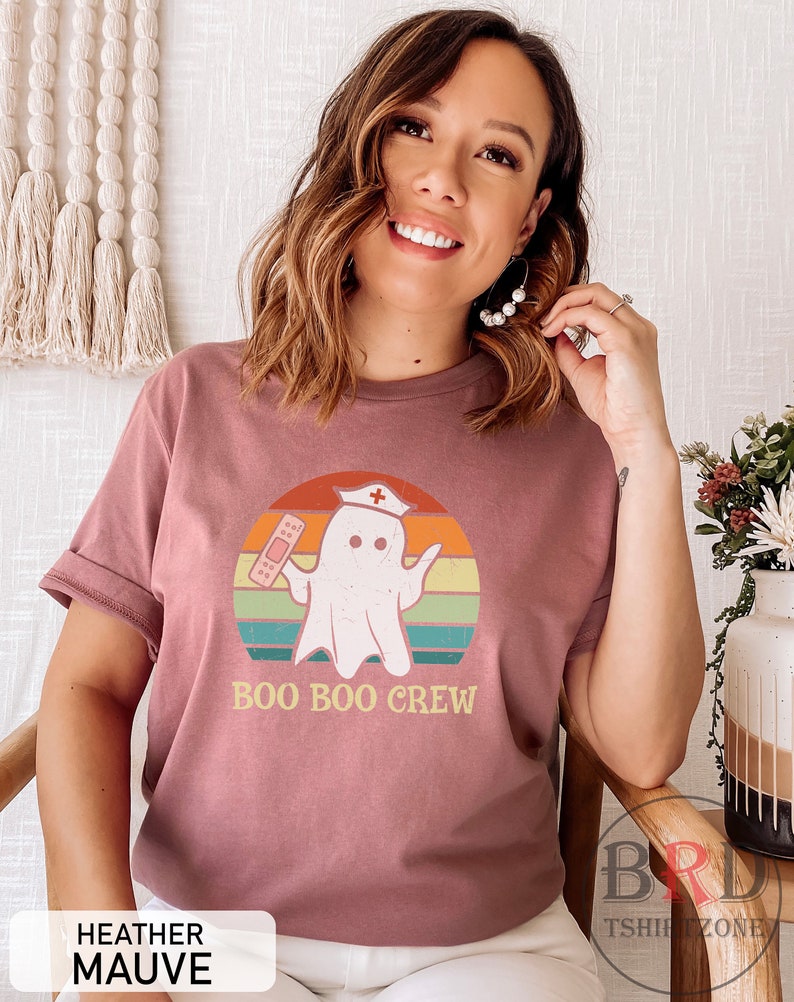 May include: Heather mauve t-shirt with a graphic of a ghost wearing a nurse's hat and holding a bandage, set against a retro sunset design. The text "BOO BOO CREW" is below the graphic. The shirt has rolled-up sleeves.