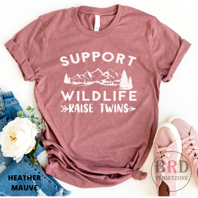 Puede incluir: Una camiseta de color malva con el texto "Support Wildlife - Raise Twins" en blanco. El texto est&aacute; rodeado de un gr&aacute;fico de monta&ntilde;as, &aacute;rboles y un r&iacute;o.