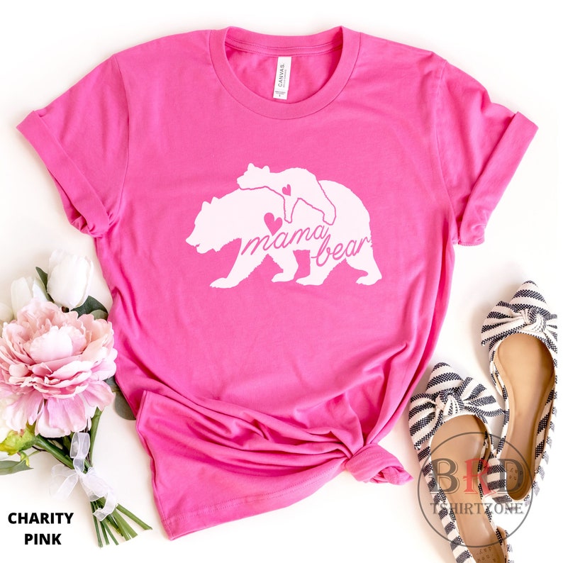 May include: A bright pink t-shirt with a white graphic of a mama bear and cub, with the words "mama bear" in script. The shirt is folded and has the words "Charity Pink" at the bottom.