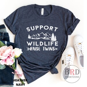 Puede incluir: Una camiseta azul marino con texto blanco que dice "Apoya la vida silvestre - Cr&iacute;a gemelos". El texto est&aacute; acompa&ntilde;ado de un gr&aacute;fico de una cordillera y un pino.