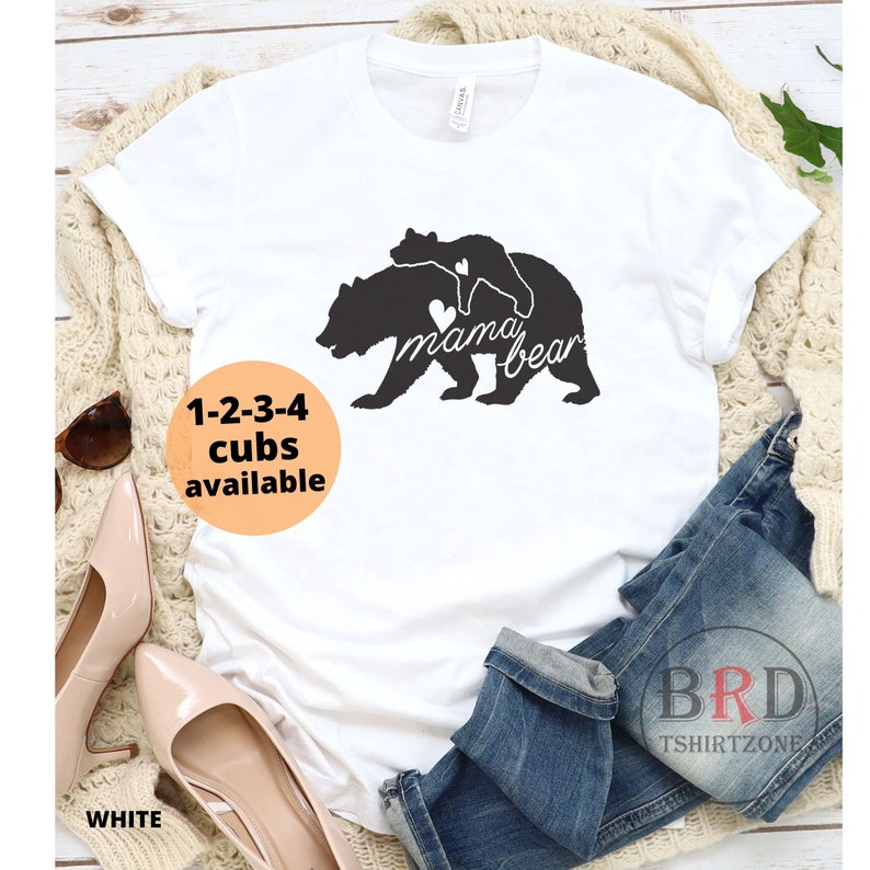 May include: White t-shirt with a black graphic of a mama bear and cub, with the text "mama bear" and a heart. The shirt also has the text "1-2-3-4 cubs available" in an orange circle. The word "WHITE" is at the bottom.