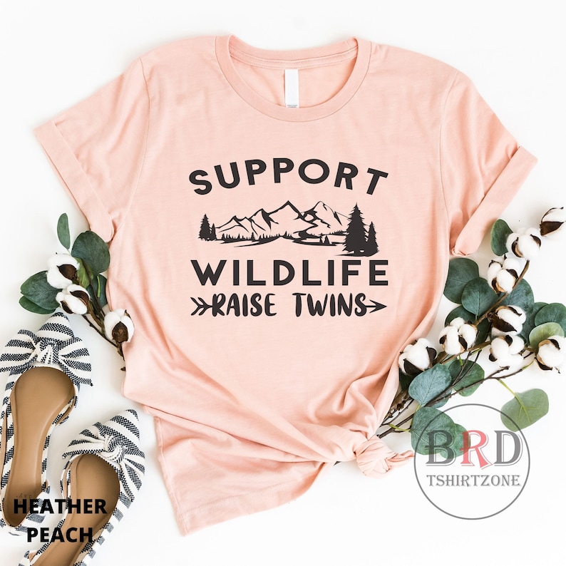 Puede incluir: Una camiseta rosa con el texto "Support Wildlife Raise Twins" en negro. El texto est&aacute; rodeado por un gr&aacute;fico de monta&ntilde;as, &aacute;rboles y un lago.