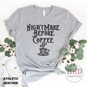 May include: Athletic heather gray t-shirt with the text "NIGHTMARE BEFORE COFFEE" in black. The design includes a coffee cup with a skull face. The shirt is styled with white sneakers and decorative greenery.