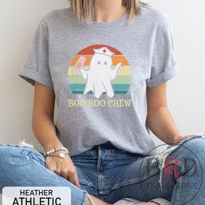 May include: Heather gray t-shirt with a graphic of a ghost wearing a nurse's cap and holding a bandage, set against a retro sunset design. The text "BOO BOO CREW" is below the graphic. The shirt also has the text "HEATHER ATHLETIC".