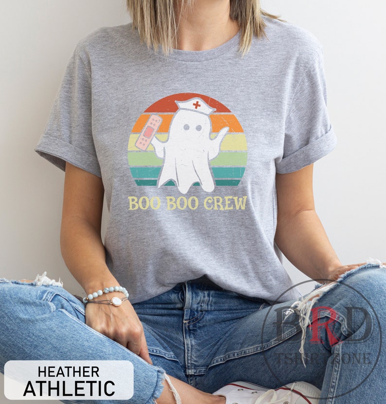 May include: Heather gray t-shirt with a graphic of a ghost wearing a nurse's cap and holding a bandage, set against a retro sunset design. The text "BOO BOO CREW" is below the graphic. The shirt also has the text "HEATHER ATHLETIC".