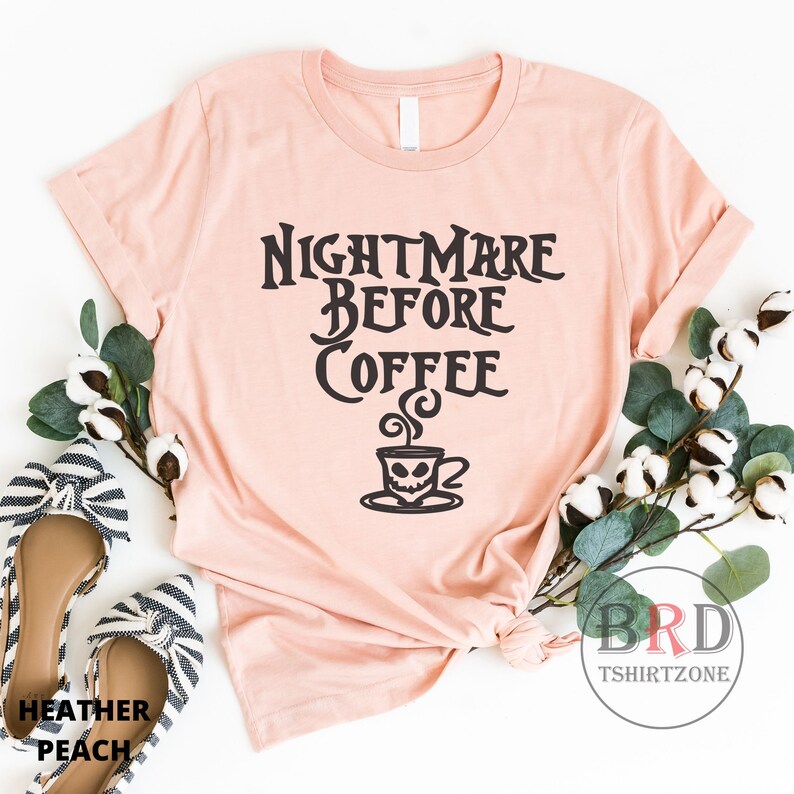 May include: A heather peach t-shirt with the text "NIGHTMARE BEFORE COFFEE" in a gothic font. The design includes a coffee cup with a skull face. The shirt is styled with decorative greenery and a pair of striped shoes.
