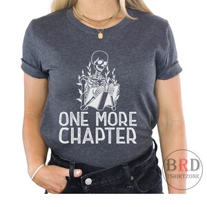 May include: A dark gray t-shirt with a white skeleton reading a book. The text "ONE MORE CHAPTER" is printed below the skeleton.