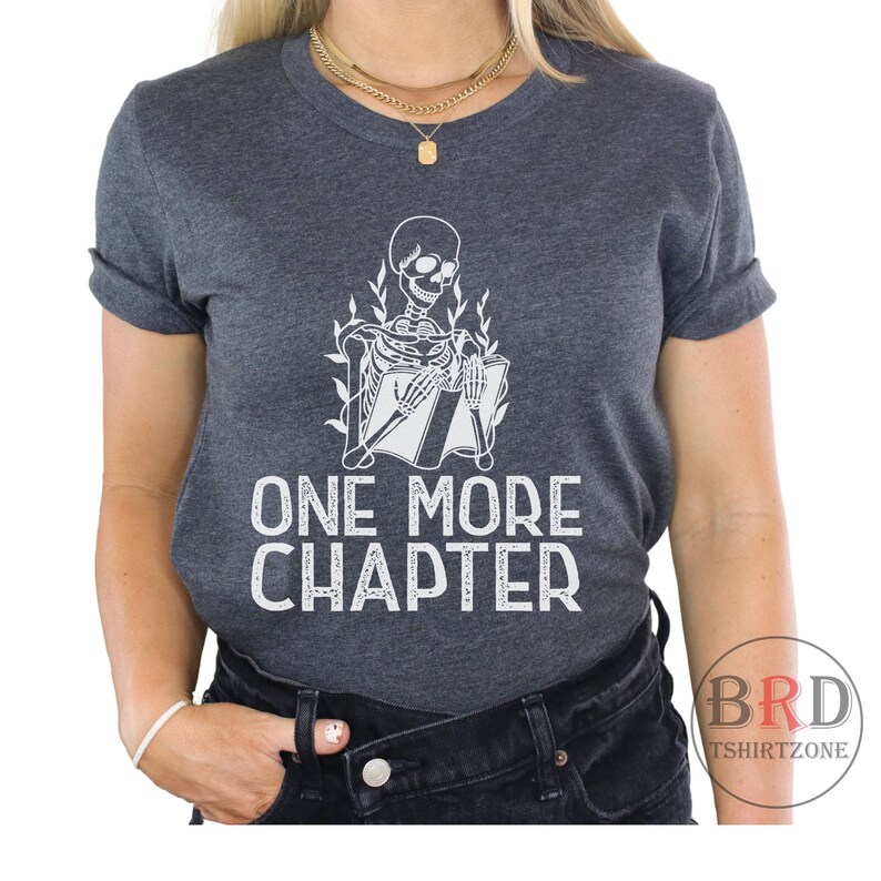 May include: A dark gray t-shirt with a white skeleton reading a book. The text "ONE MORE CHAPTER" is printed below the skeleton.