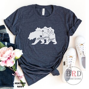 May include: Heather navy t-shirt with a white graphic of a bear and three cubs, with the words "mama bear" in script. The shirt is styled with rolled sleeves, jeans, flowers, and white sneakers.