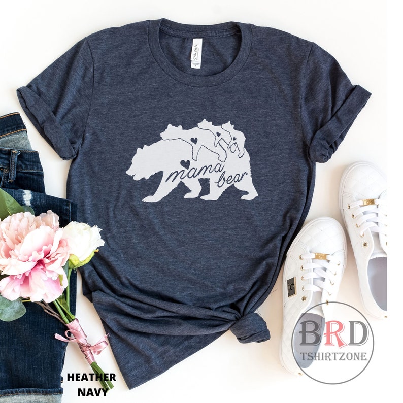 May include: Heather navy t-shirt with a white graphic of a bear and three cubs, with the words "mama bear" in script. The shirt is styled with rolled sleeves, jeans, flowers, and white sneakers.