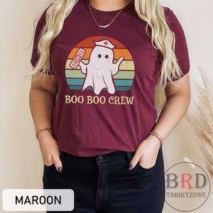 May include: Maroon t-shirt featuring a cartoon ghost wearing a nurse's cap and holding a bandage, set against a retro sunset design. The text "BOO BOO CREW" is below the graphic. The shirt is from BRD TSHIRTZONE.