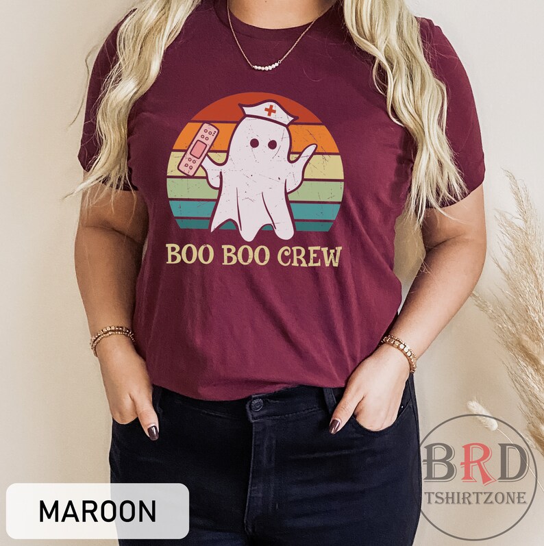 May include: Maroon t-shirt featuring a cartoon ghost wearing a nurse's cap and holding a bandage, set against a retro sunset design. The text "BOO BOO CREW" is below the graphic. The shirt is from BRD TSHIRTZONE.