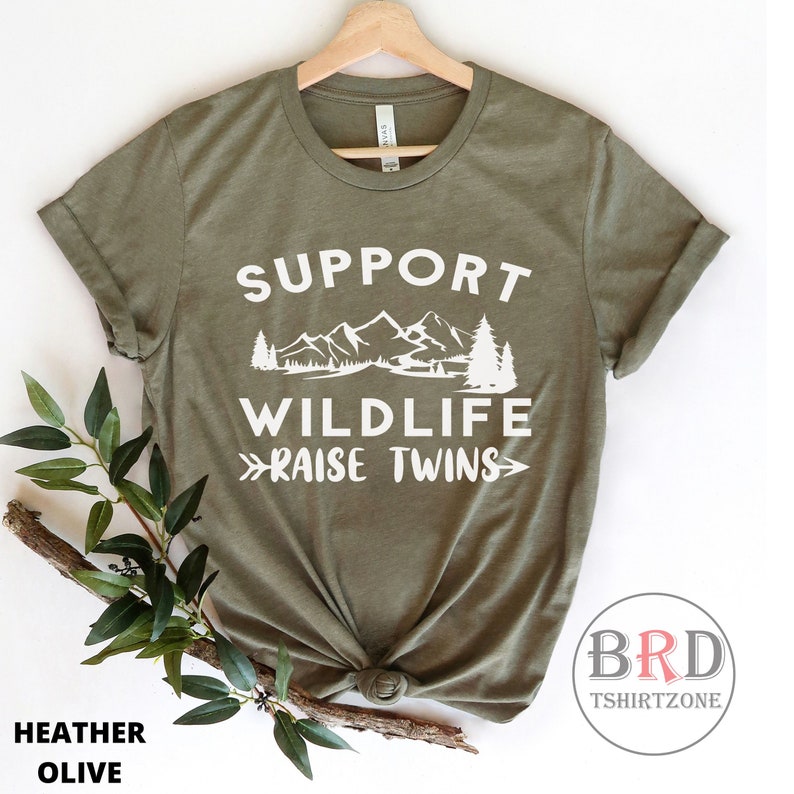 Puede incluir: Una camiseta verde con texto blanco que dice "Support Wildlife Raise Twins". El texto est&aacute; rodeado de un gr&aacute;fico de monta&ntilde;as y &aacute;rboles.