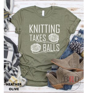 May include: A green t-shirt with the text "Knitting Takes Balls" in white lettering. Two white yarn balls are pictured below the text.