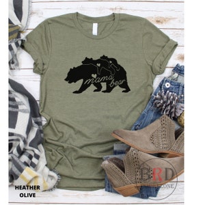 May include: Heather olive green t-shirt with a black graphic of a mama bear and two cubs. The words "mama bear" are written in a script font. The shirt is displayed with jeans and boots.