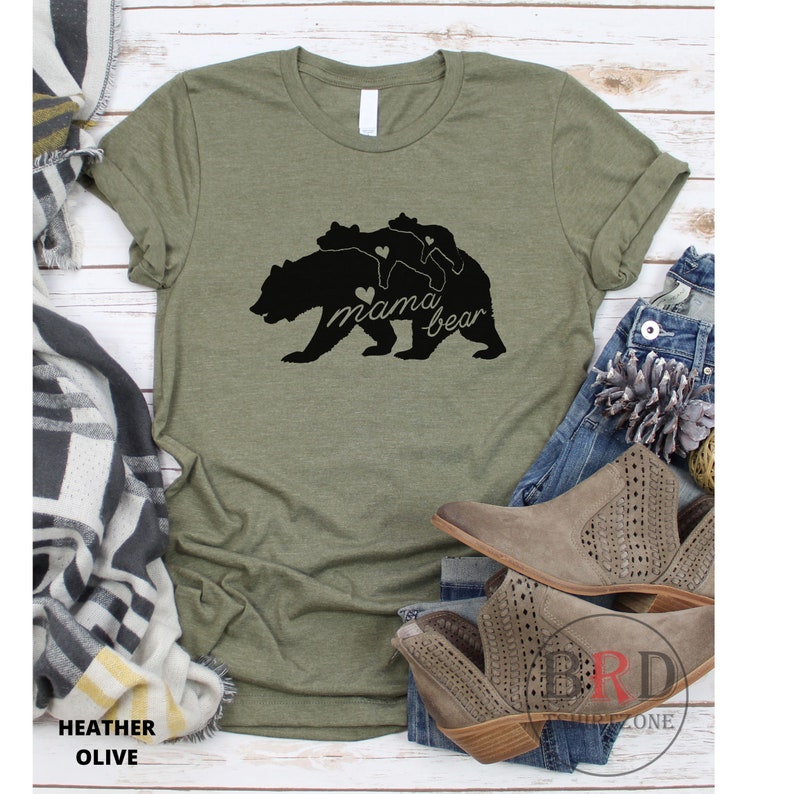 May include: Heather olive green t-shirt with a black graphic of a mama bear and two cubs. The words "mama bear" are written in a script font. The shirt is displayed with jeans and boots.