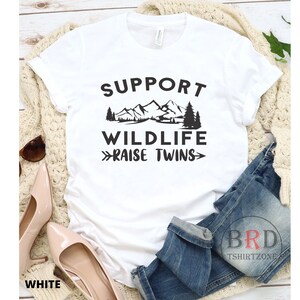 Puede incluir: Camiseta blanca con texto negro que dice "Support Wildlife Raise Twins" con un gr&aacute;fico de monta&ntilde;as y &aacute;rboles.