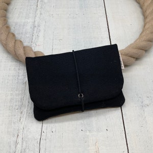 Könnte beinhalten: Eine schwarze Stofftasche mit Kordelzug und einem Metallring. Die Tasche liegt auf einer weißen Holzoberfläche.
