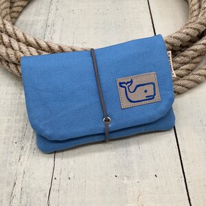 Könnte beinhalten: Eine blaue Stofftasche mit einem grauen Kordelverschluss und einem braunen Lederflicken mit einem blauen Wal-Logo. Die Tasche liegt auf einer Holzoberfläche.