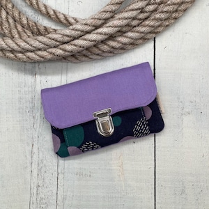 Può includere: Un piccolo portafoglio rettangolare con patta viola e corpo blu navy a fantasia con cerchi verdi e viola. Il portafoglio ha una chiusura argentata ed è appoggiato su uno sfondo di legno bianco con una corda.