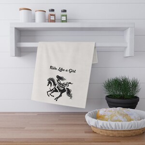 May include: A cream-colored rectangular kitchen towel hanging from a white shelf. The towel features a black graphic print of a person riding a horse with flowing hair and the black text Ride Like a Girl printed above the illustration.