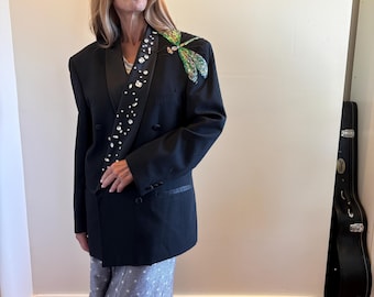 Könnte beinhalten: Ein schwarzer zweireihiger Blazer mit einem Satin-Revers, das mit klaren Kristallen verziert ist, und einer großen, bunten, schillernden Libellen-Applikation auf der linken Schulter. Der Blazer wird über einem grauen Rock mit weißen Sternen getragen.