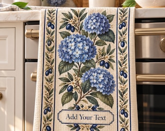 Peut inclure: Un torchon de cuisine en tissage gaufré couleur crème orné d'une illustration botanique centrale de fleurs d'hortensia bleues et de fruits ovales bleu foncé. Le motif inclut des bordures latérales et un cadre avec le texte Add Your Text.