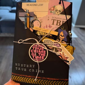 May include: A black envelope-style folder labeled MYSTERY TRUE CRIME containing a reading list, a yellow pen, a skull keychain, tea, and stickers. A circular sticker reads TRUE CRIME ALL THE TIME. The package is tied with white string.