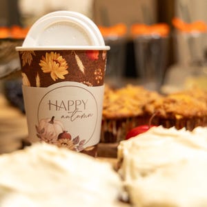 May include: A brown paper coffee cup with a white plastic lid featuring a pattern of orange flowers, wheat, and pumpkins. A beige sleeve wraps around the center with the text "HAPPY autumn" printed in black, accompanied by a small pumpkin illustration.