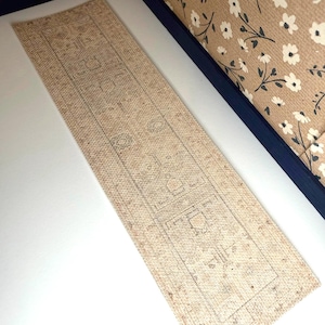 May include: A rectangular runner rug featuring a light beige base with a faded, intricate geometric pattern in muted gray tones. The rug has a textured, woven appearance and is positioned on a flat surface next to a wall with floral wallpaper.
