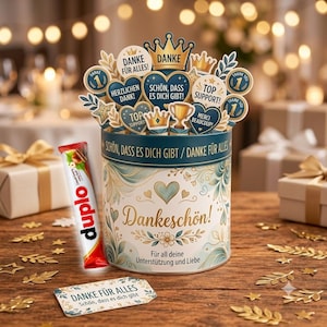 May include: A cylindrical gift tin with a white and teal floral design, featuring the text Dankeschön and Für all deine Unterstützung und Liebe. It is topped with decorative cutouts of hearts, crowns, and trophies, and accompanied by a Duplo chocolate bar and a rectangular tag reading Danke für alles.