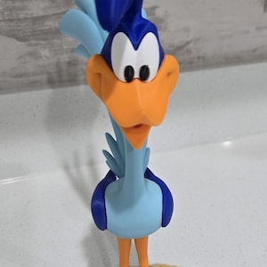 May include: A 3D printed figurine of the Road Runner character, featuring a tall blue crest, a bright orange beak, and a light blue body. The bird stands on a circular tan base with a textured, wavy surface resembling desert sand.