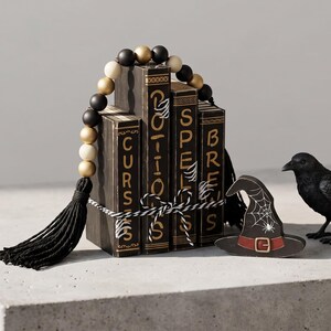 May include: A stack of four dark books labeled Curses, Potions, Spells, and Brews, tied with black and white twine. A string of black, gold, and cream beads with black tassels arches over the books. A black crow and a witch hat are nearby.