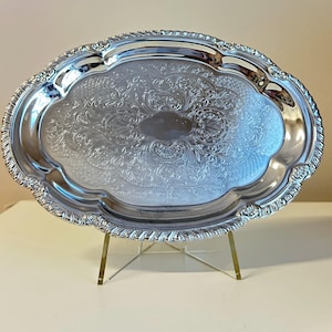 May include: An oval silver-toned serving tray featuring an intricate embossed floral and scrollwork pattern across its surface. The tray has a scalloped edge with a decorative rope-like border and a smooth, polished center area.