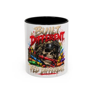 May include: A white ceramic mug with a black interior featuring a graphic of a skull wearing a tactical helmet and goggles, biting a red crayon. The design includes colorful scattered crayons and the text BUILT DIFFERENT and FED DIFFERENT.
