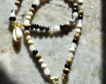 Puede incluir: Un collar de cuentas que presenta una variedad de pequeñas cuentas redondas en tonos negro, blanco, marrón y gris translúcido. El collar incluye dos cierres de metal dorado, cada uno adornado con un colgante de concha cauri blanca.