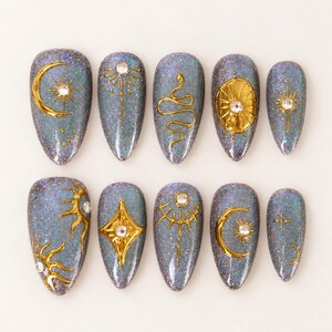 Puede incluir: Un juego de diez uñas postizas en forma de almendra con un acabado de purpurina holográfica gris oscuro. Cada uña presenta motivos celestiales dorados en relieve, incluyendo lunas, soles, estrellas y una serpiente, con pequeños brillantes blancos.