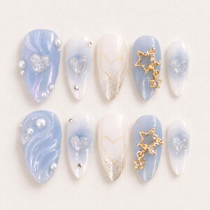 Puede incluir: Un juego de diez uñas postizas en forma de almendra con una paleta de colores azul suave y blanco. Los diseños incluyen detalles de corazones iridiscentes, dijes de estrellas doradas con cristales, perlas blancas, degradados de purpurina y líneas doradas.