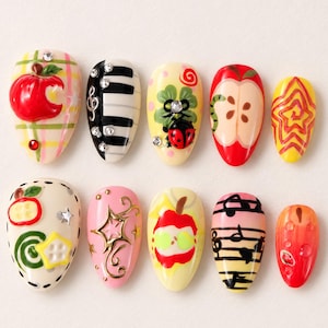 Puede incluir: Un juego de diez uñas postizas en forma de almendra con diversos diseños que incluyen una manzana roja, teclas de piano con pedrería, una mariquita sobre un trébol, rodajas de manzana, estrellas amarillas, notas musicales y detalles dorados sobre bases rosa y crema.