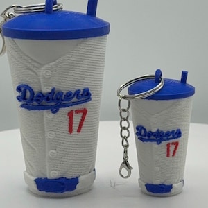 May include: Two white and blue cup-shaped keychains featuring a textured baseball jersey design. Each cup has a blue lid with a straw, the blue text Dodgers, and the red number 17 printed on the front. A metal keyring is attached to each lid.